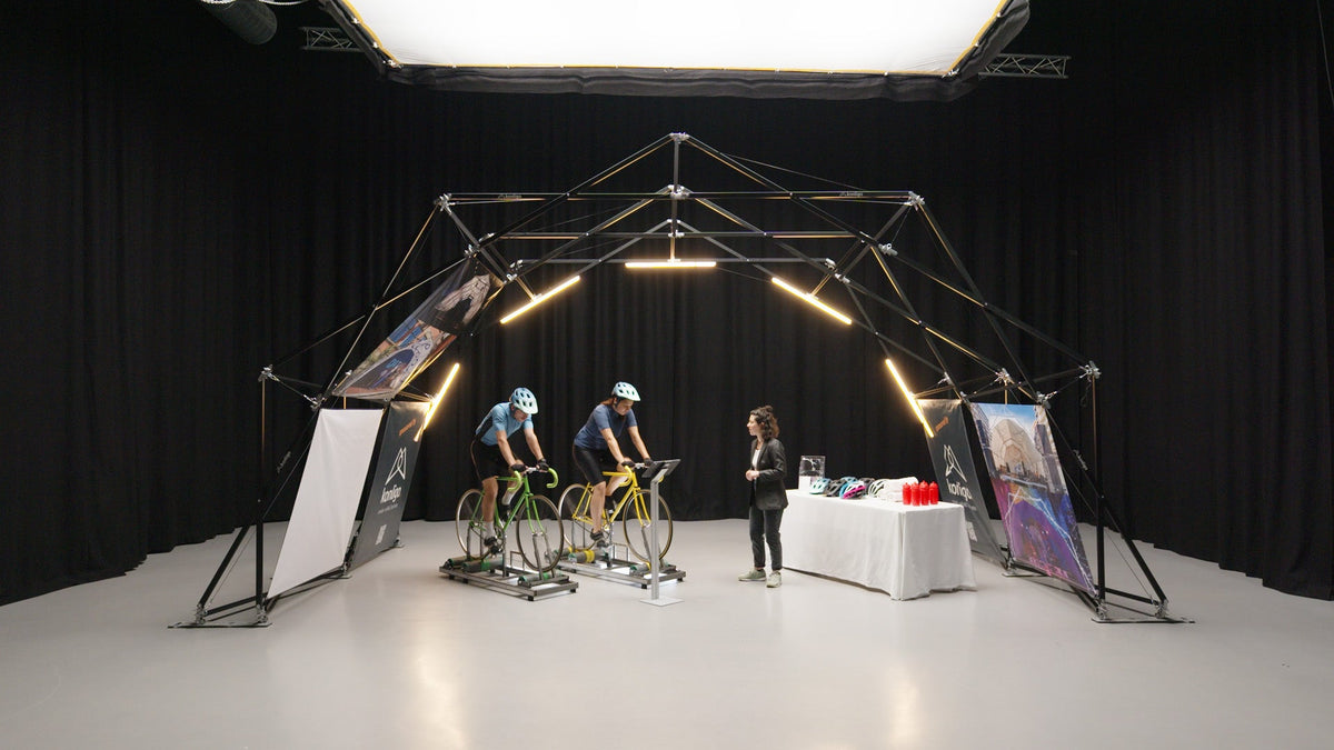 Indoor setup of Konligo event structure featuring two cyclists on stationary bikes and a display table with helmets and water bottles. Now available on MTN Shop US.
 - imagen 4